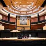 Interno di un teatro storico con platea, palco illuminato e sedute rosse, pronto per un grande evento.