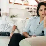Una coppia seduta in salotto ha una conversazione seria, con documenti e un calendario visibili sul tavolo.