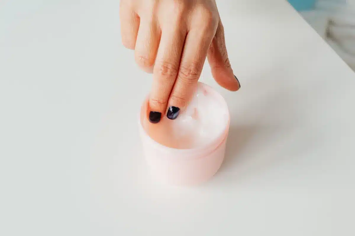 Persona che applica crema idratante al viso con le mani, circondata da prodotti skincare