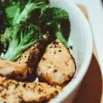 Mani che tengono un frullato proteico e una bottiglia d'acqua accanto a un piatto con pollo e verdure