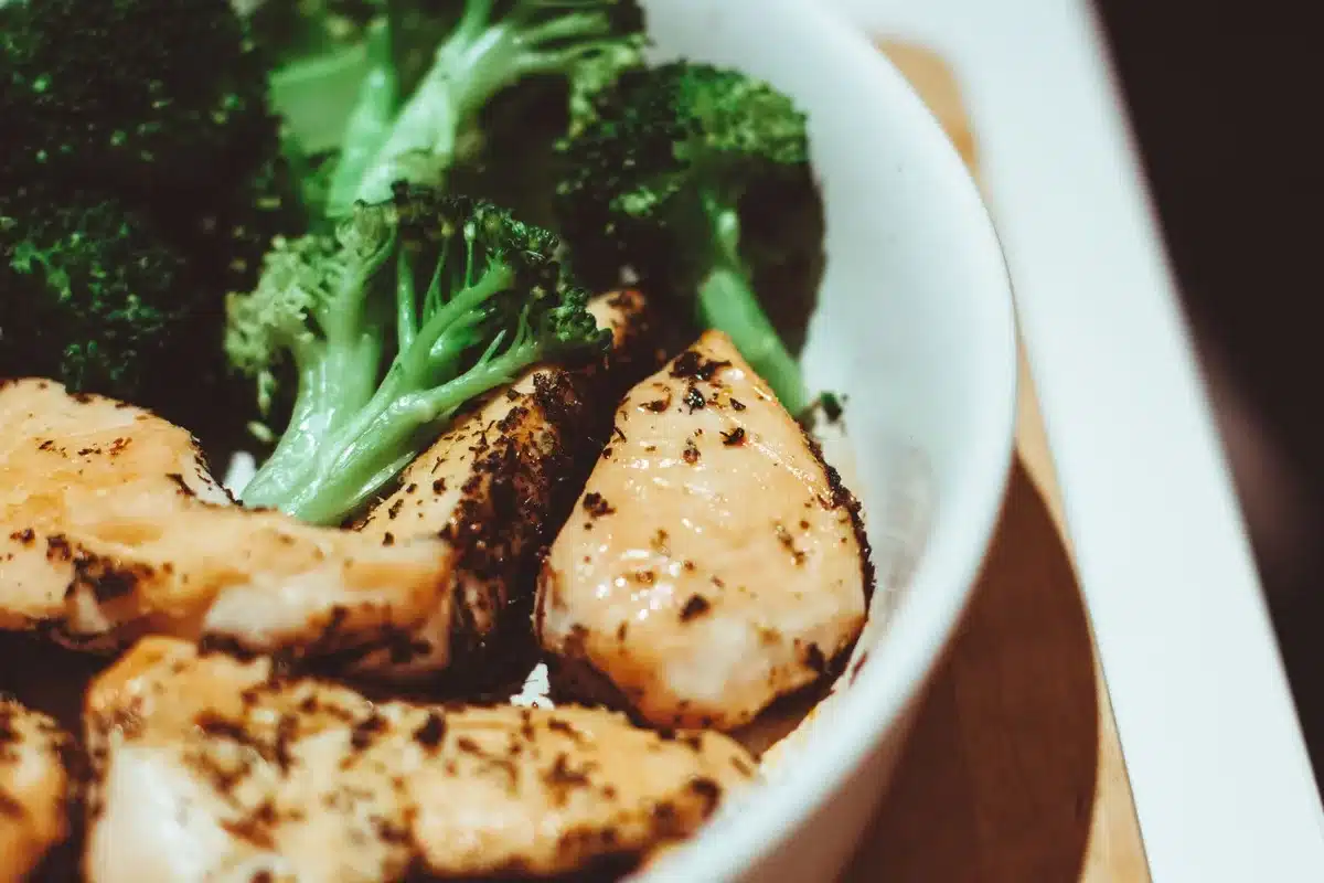 Mani che tengono un frullato proteico e una bottiglia d'acqua accanto a un piatto con pollo e verdure