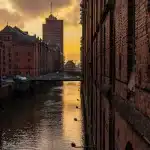 Passanti camminano lungo il Naviglio di Milano, tra edifici storici e l'arco di Porta Ticinese al tramonto