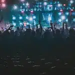 Pubblico anonimo in platea e palco con luci calde ed elementi scenici astratti